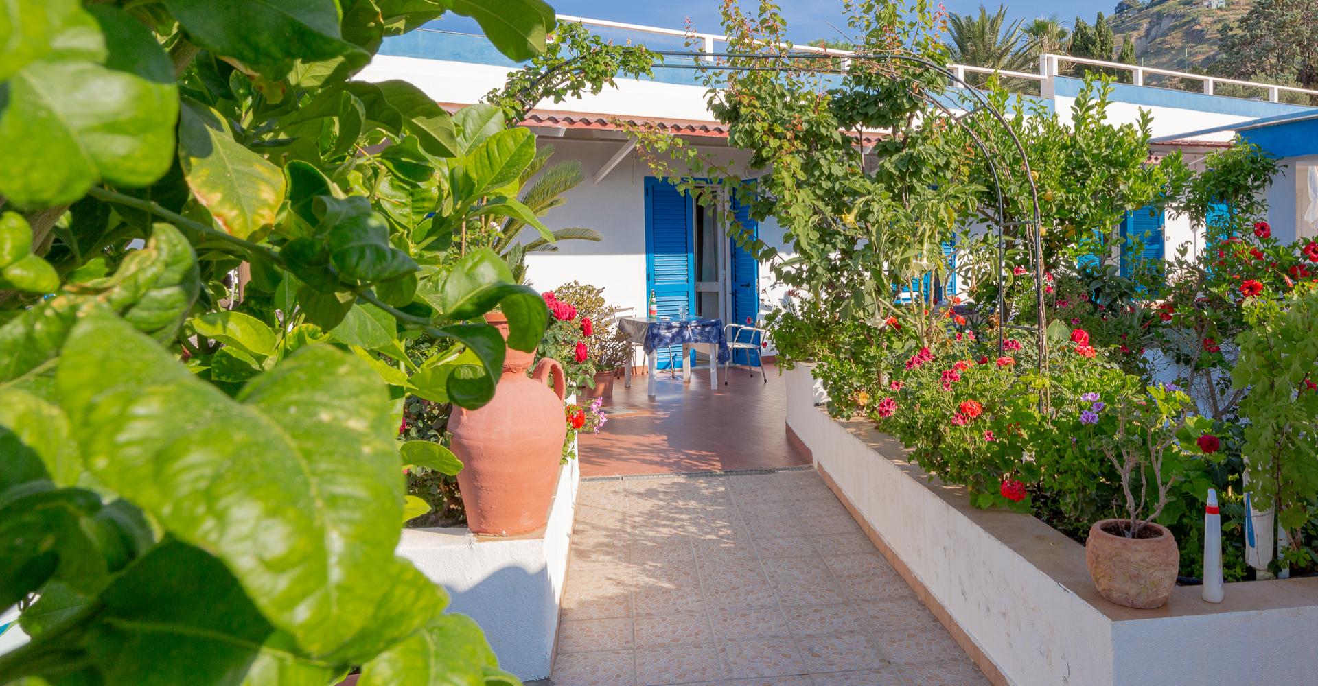 Giardino fiorito con vasi e sedie blu davanti a una casa.